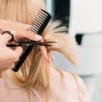 Fryzjer stylizuje długie blond włosy klientki za pomocą grzebienia i nożyczek. Profesjonalna usługa.