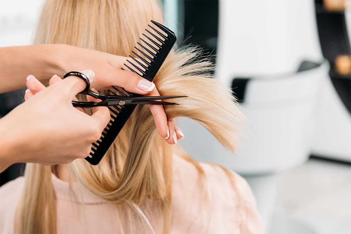 Fryzjer stylizuje długie blond włosy klientki za pomocą grzebienia i nożyczek. Profesjonalna usługa.