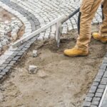 Układanie kostki brukowej w Giedlarowej - pracownik wyrównuje piasek pod szare kostki chodnikowe.